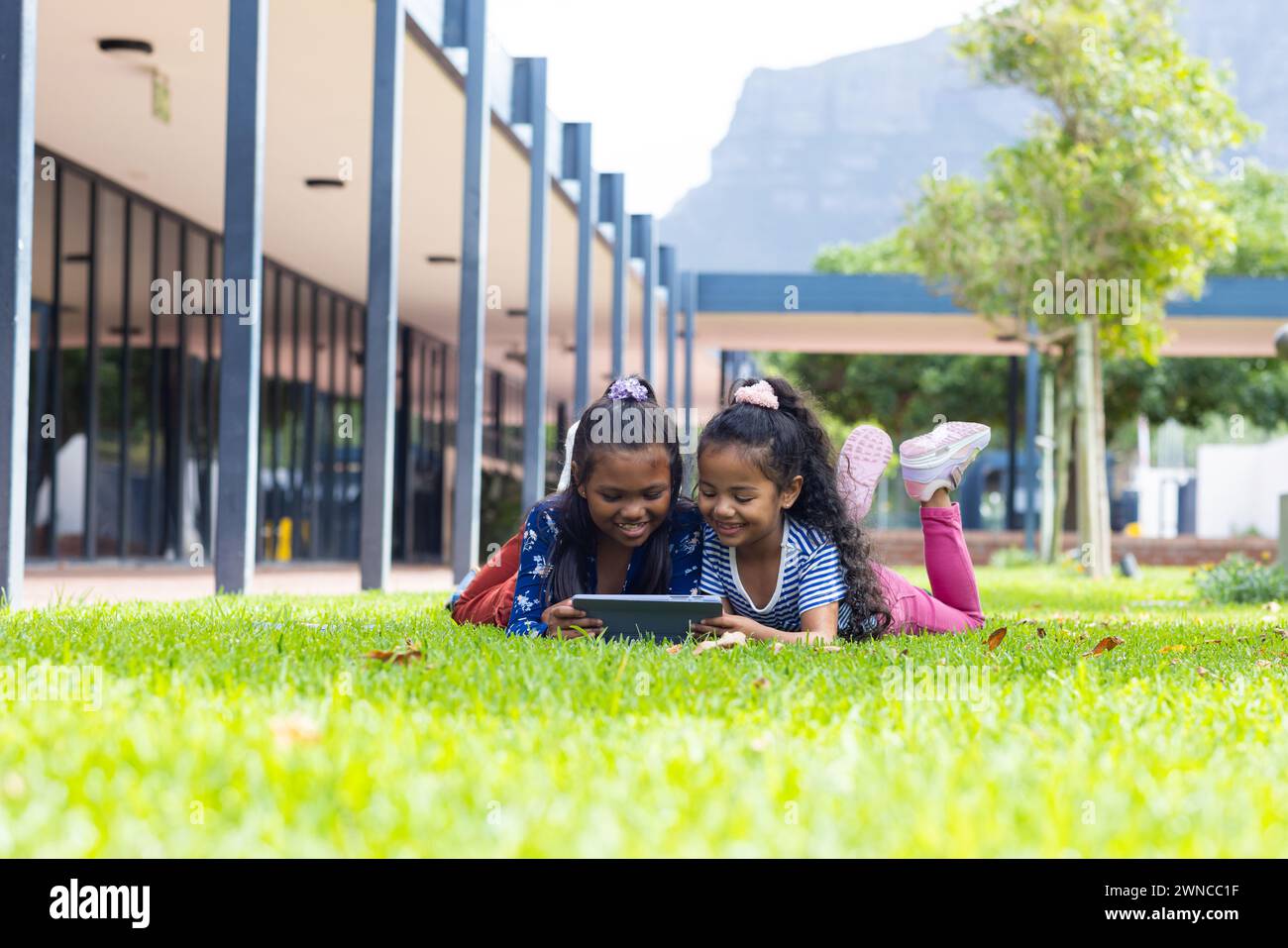 Two biracial girls are engrossed in a tablet outdoors at school, lying on the grass Stock Photo