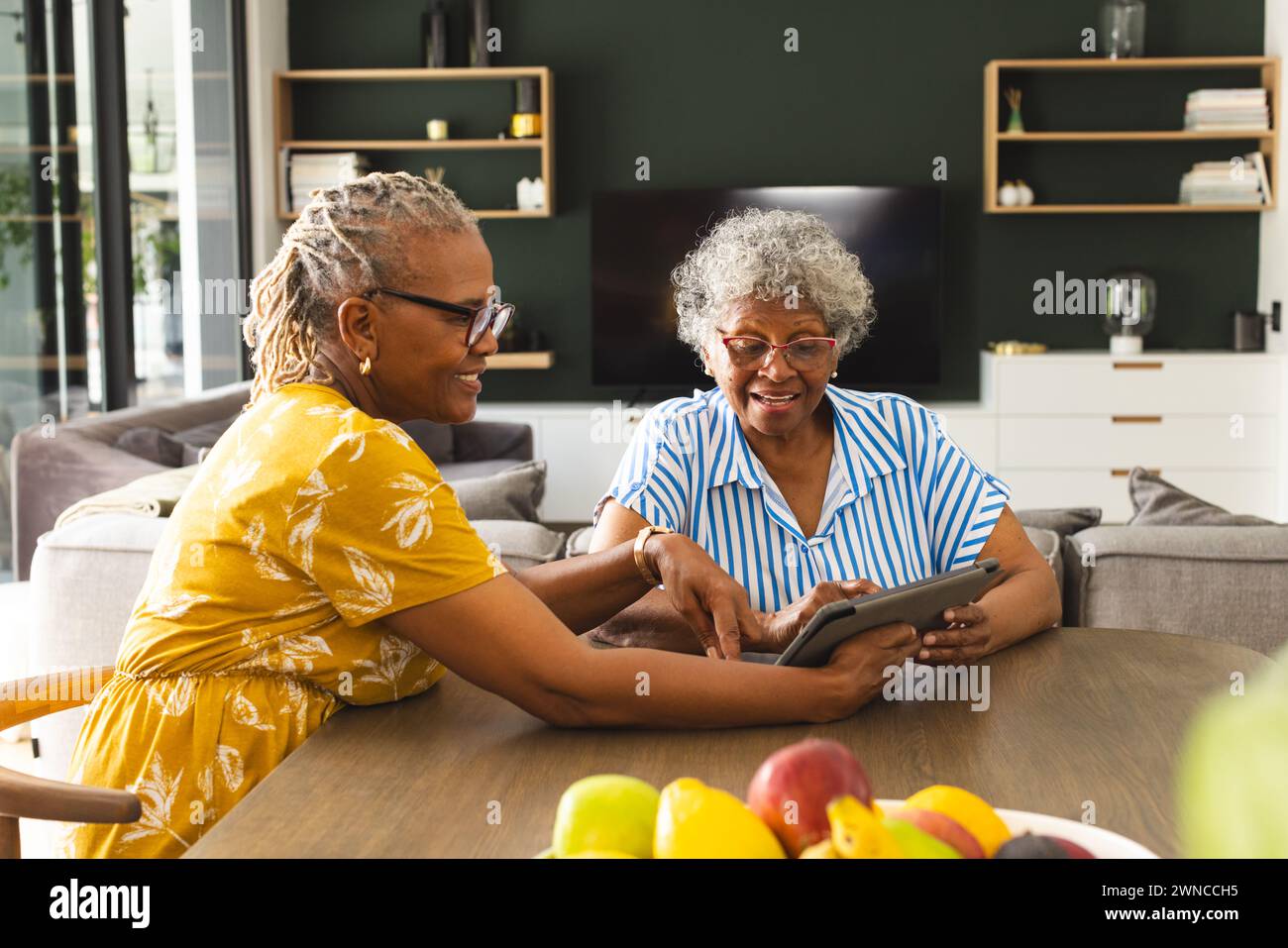 Two senior women share a tablet in a cozy, decorated living room. Stock Photo