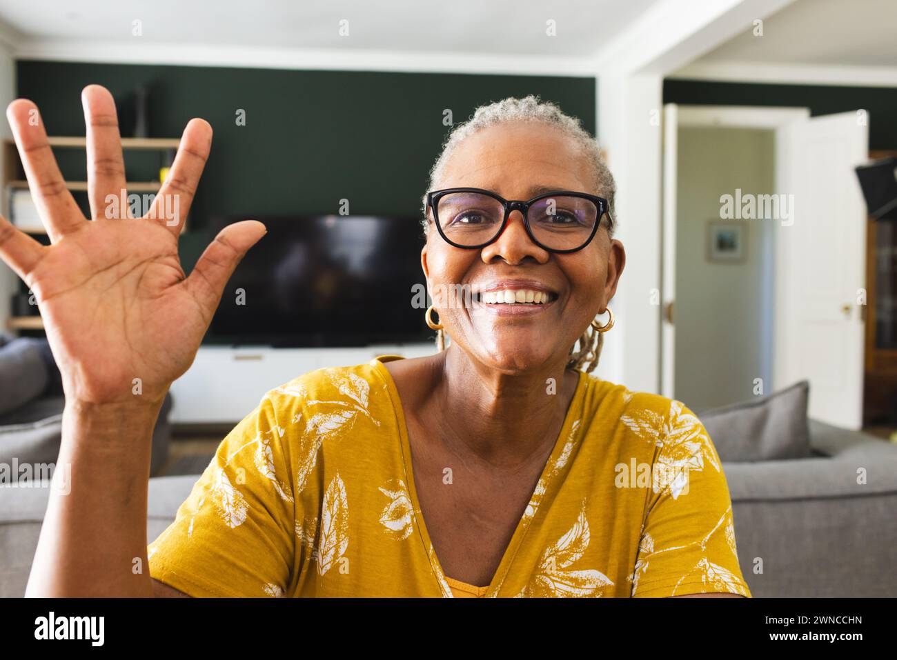 Senior African American woman waves with a bright smile, wearing glasses and a yellow top Stock Photo