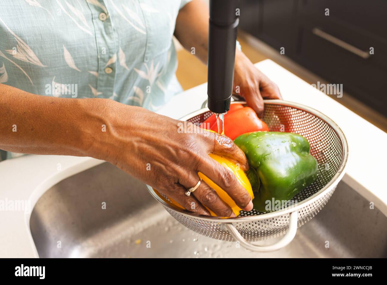 Senior biracial woman washes colorful bell peppers under a kitchen faucet Stock Photo