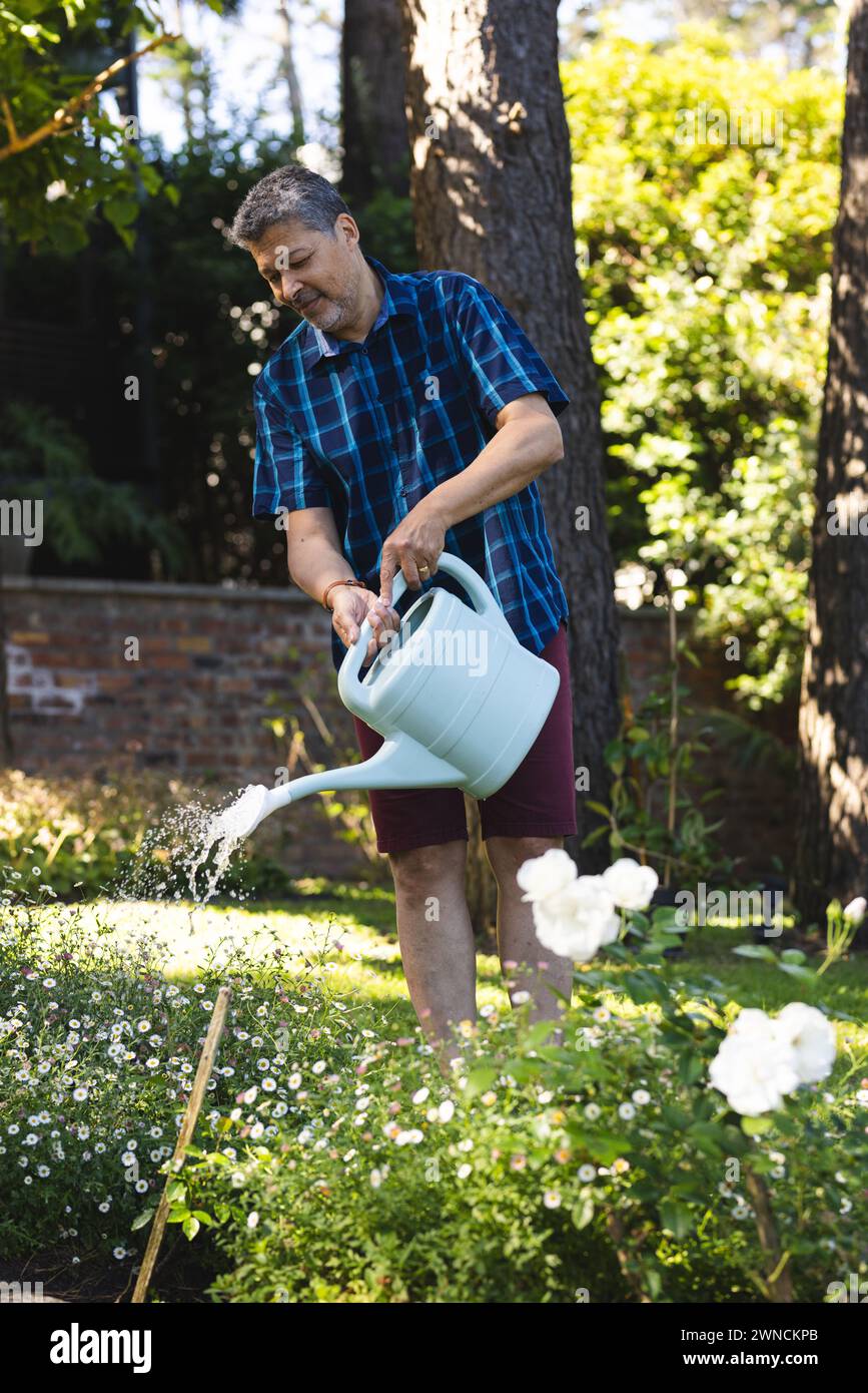 Senior biracial man waters plants in a sunny garden, wearing a blue plaid shirt and brown shorts Stock Photo
