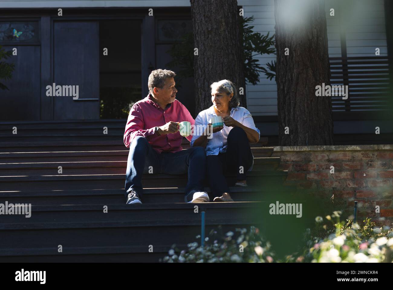 Senior biracial woman and biracial man sit on steps, enjoying a conversation with coffee Stock Photo