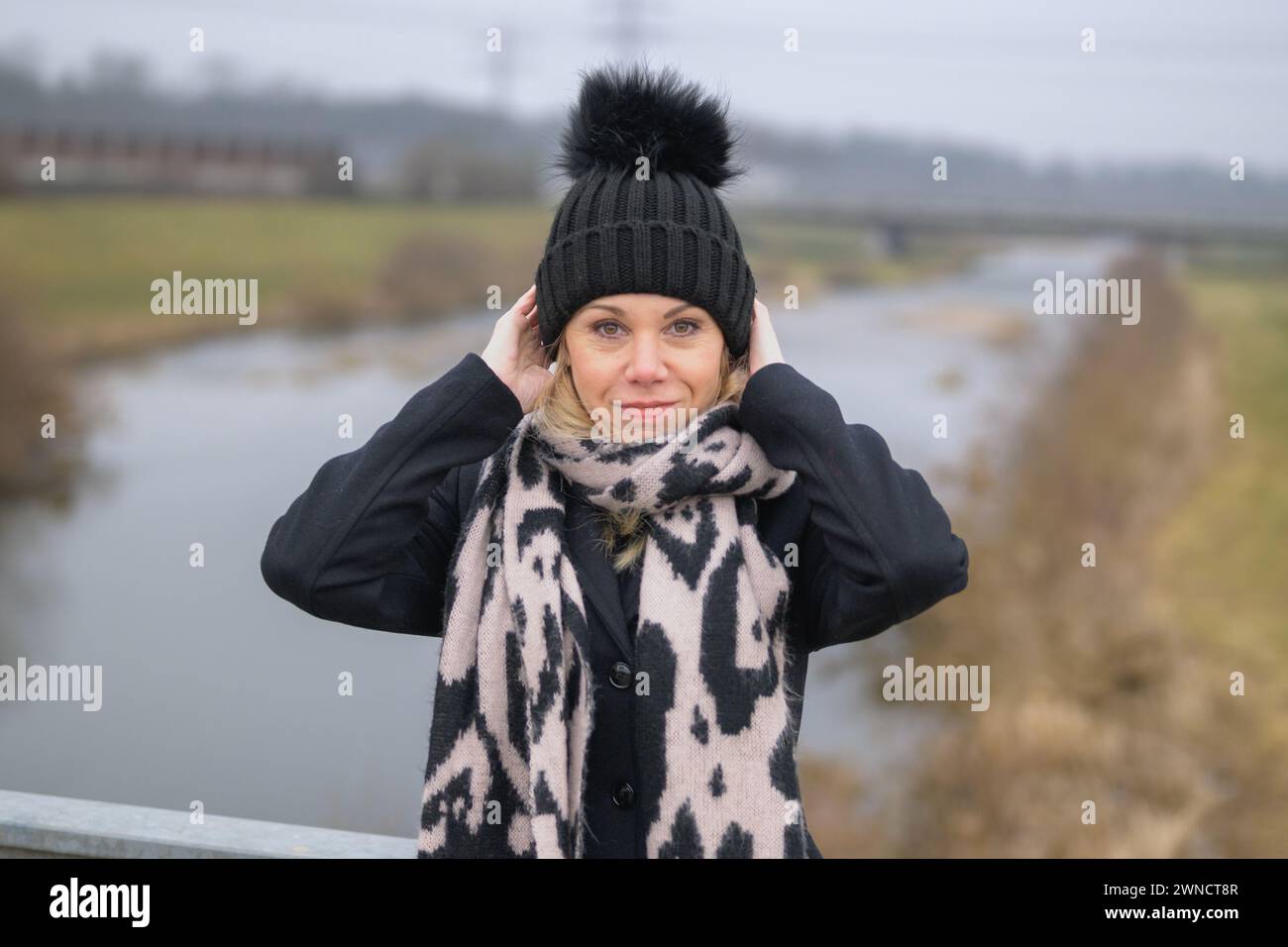 Blonde lady wearing a black coat jacket and black bobble hat holding with her hands while standing on a bridge Stock Photo