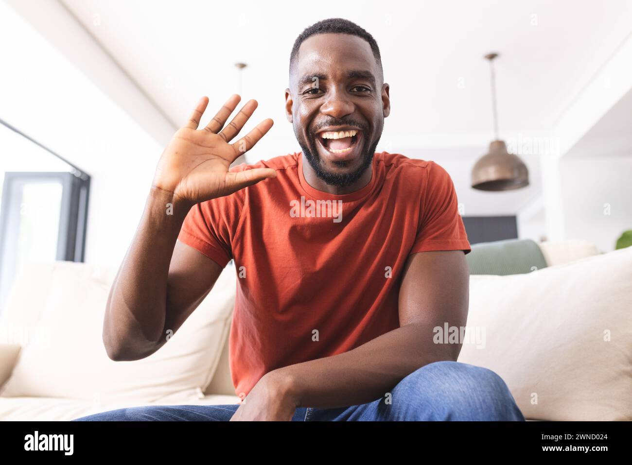 African American man in a red shirt waves with a bright smile on video call Stock Photo