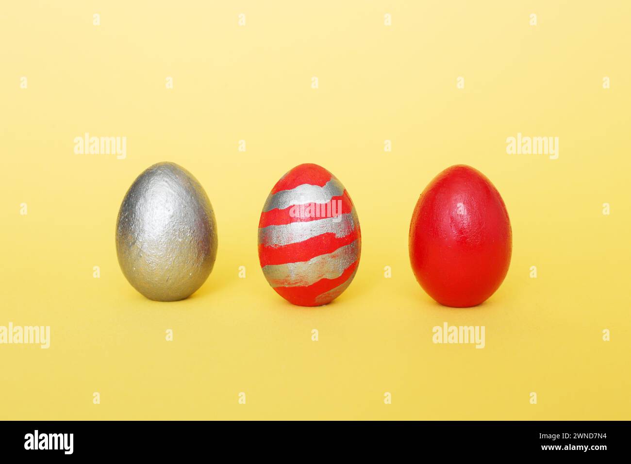 abstract composition of three multi-colored handmade eggs on a yellow pastel background. Eggs of metallic, red, red with gray stripes on a yellow pape Stock Photo
