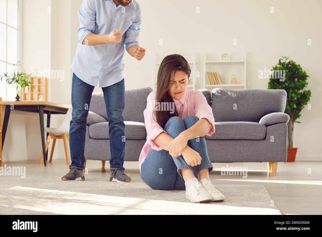 Angry man standing near upset unhappy woman sitting on floor and shouting on her at home Stock Photo