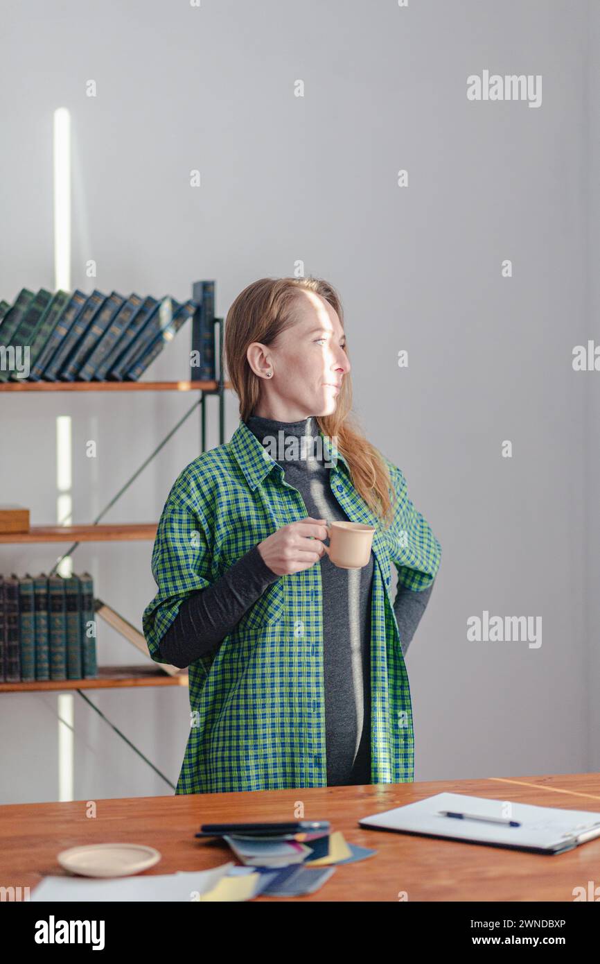 An adult young woman stands with a cup in her hand and thinks. There is a bright streak of sunlight on the face. Vertical composition. Stock Photo