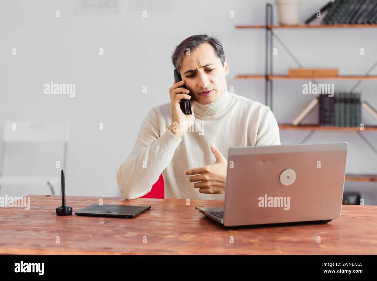 Young adult male graphic designer talking on a mobile phone in the office and working on a laptop and graphics tablet. Stock Photo