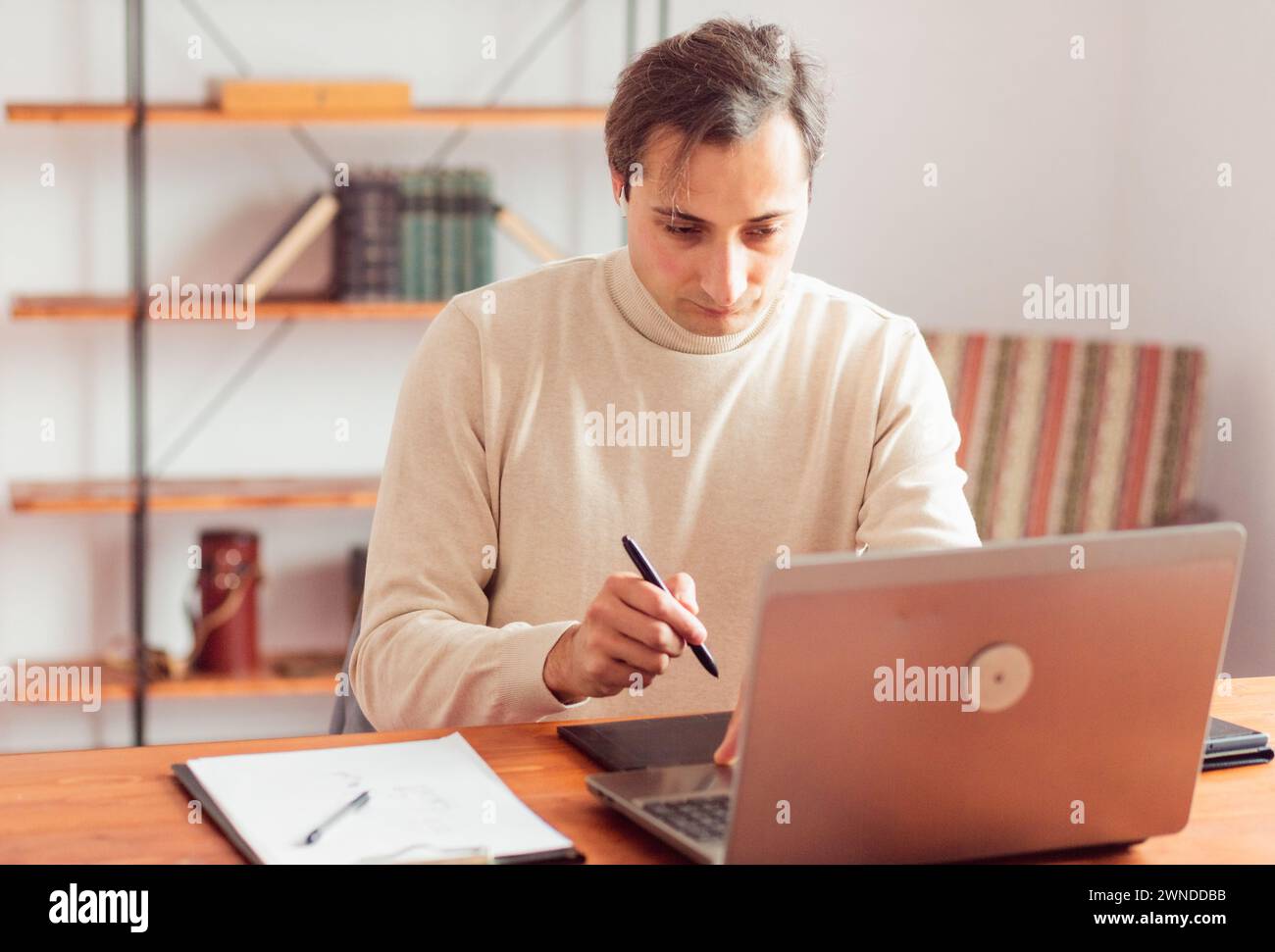 Young adult man in home office working on laptop and graphics tablet. Stock Photo