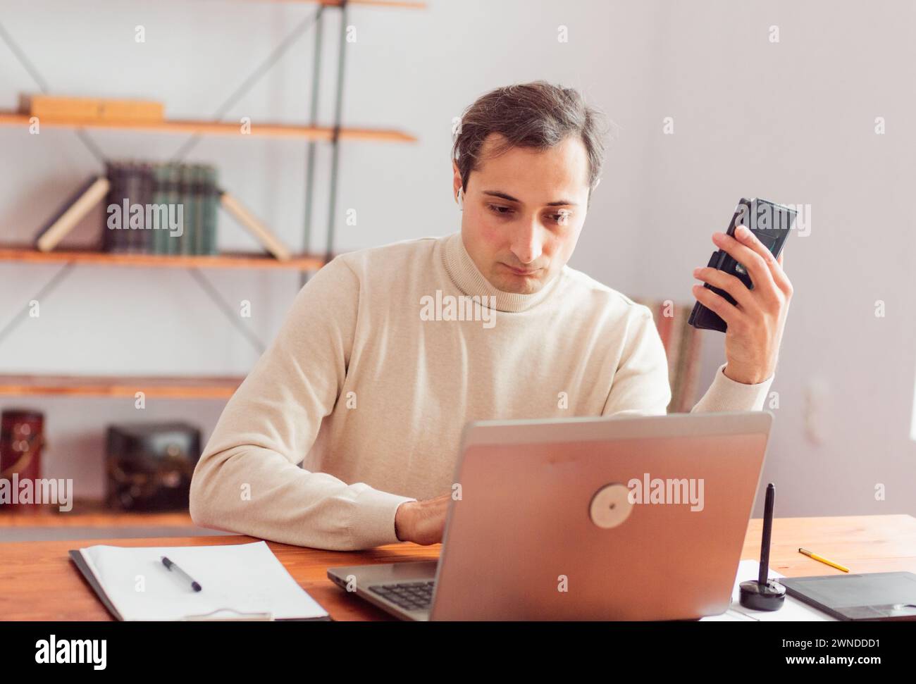 Young adult man in home office working on laptop and talking on cell phone. There are graphic electronic and paper tablets on the table. Stock Photo