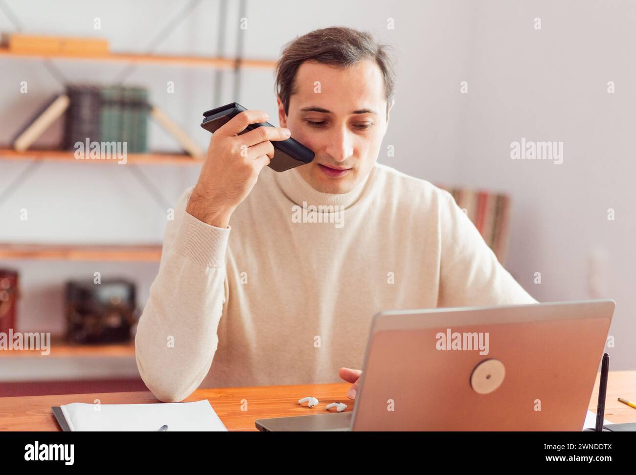 A young adult man in a home office working on a laptop talking on a cell phone. Stock Photo