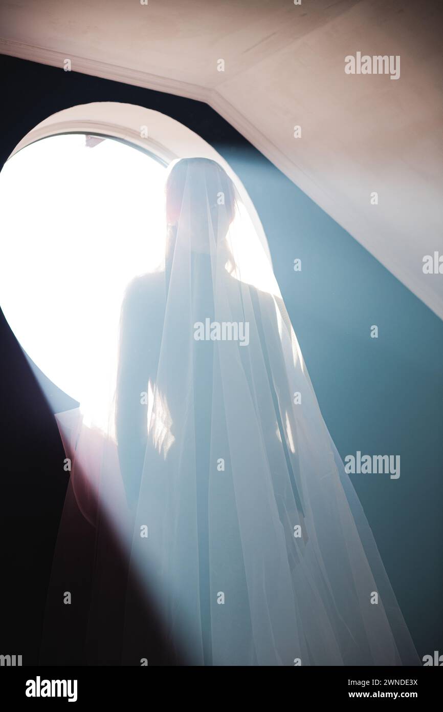 Young woman in transparent white cloth against the background of a round window in the bright rays of the sun. Hands down. Vertical composition Stock Photo