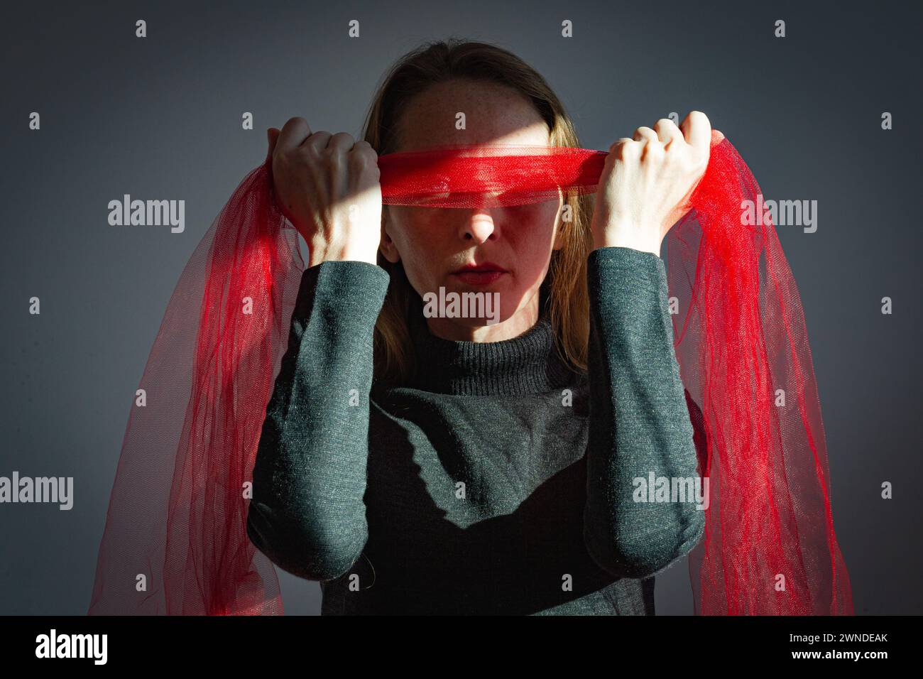Visualization of a psychological problem. An adult young woman holds a bandage made of red transparent fabric over her eyes. Stock Photo