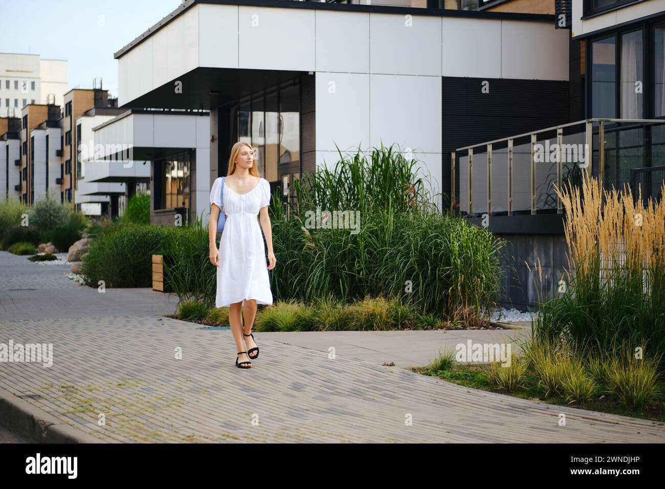 Young woman in sundress walking along the pavement in modern residential area Stock Photo