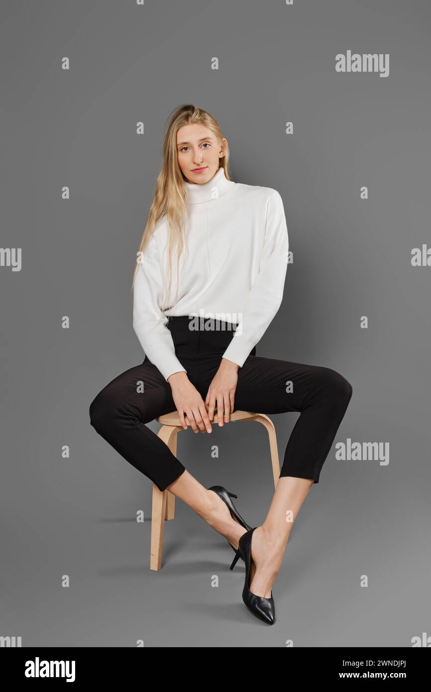 Young blonde woman in tight trousers and sweater sits on chair with legs spread to the sides Stock Photo
