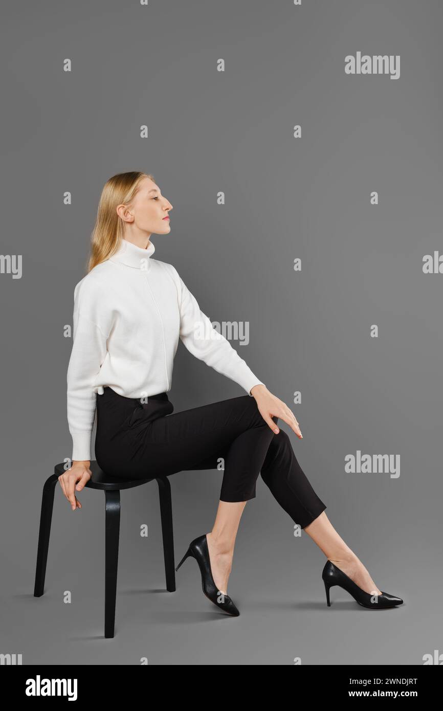 Young woman in tight trousers and white sweater sits in profile in studio Stock Photo