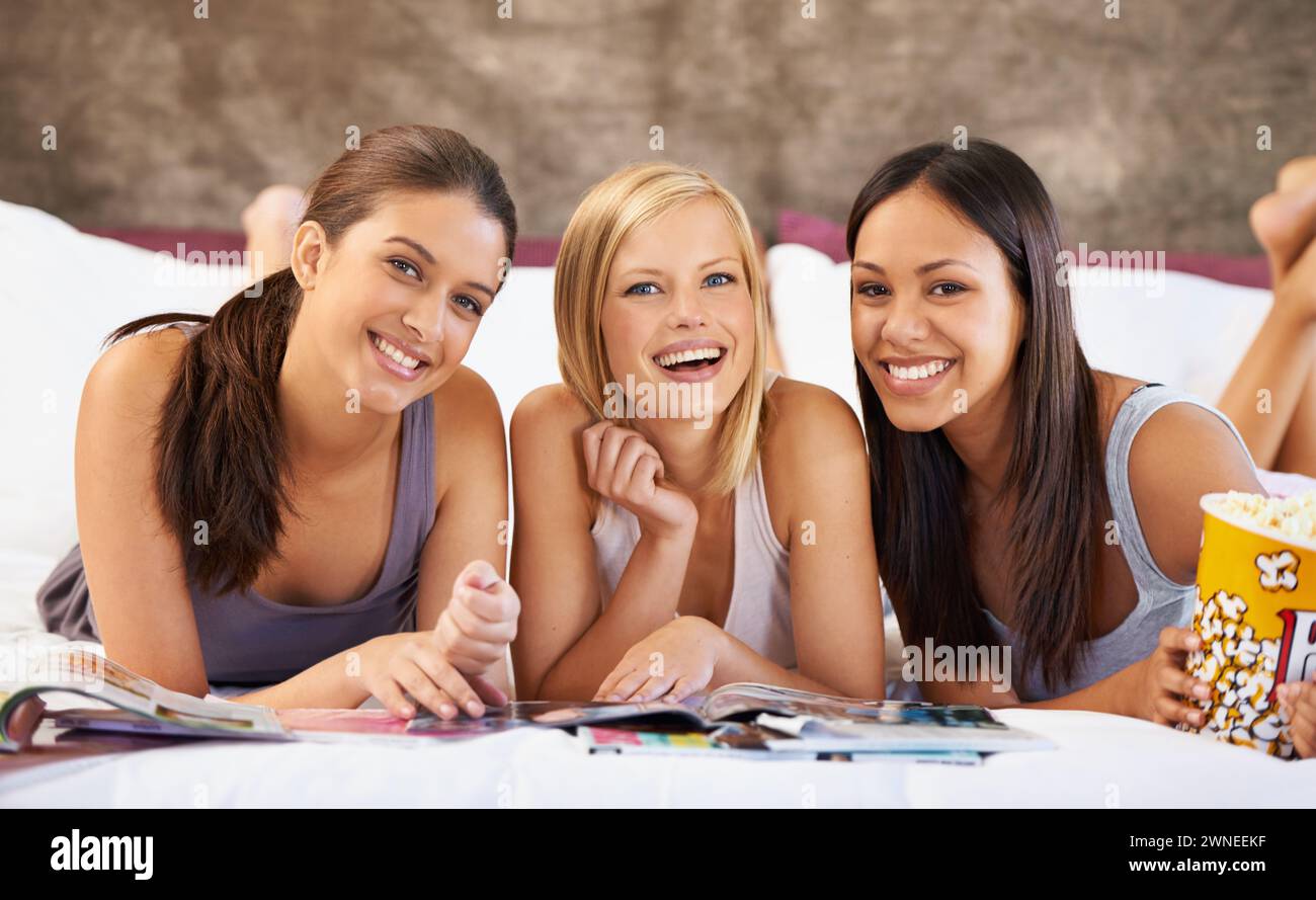 Happy friends, portrait and girls reading magazine in bedroom, food and eating popcorn snack together at party. Smile, women and group with journal in Stock Photo