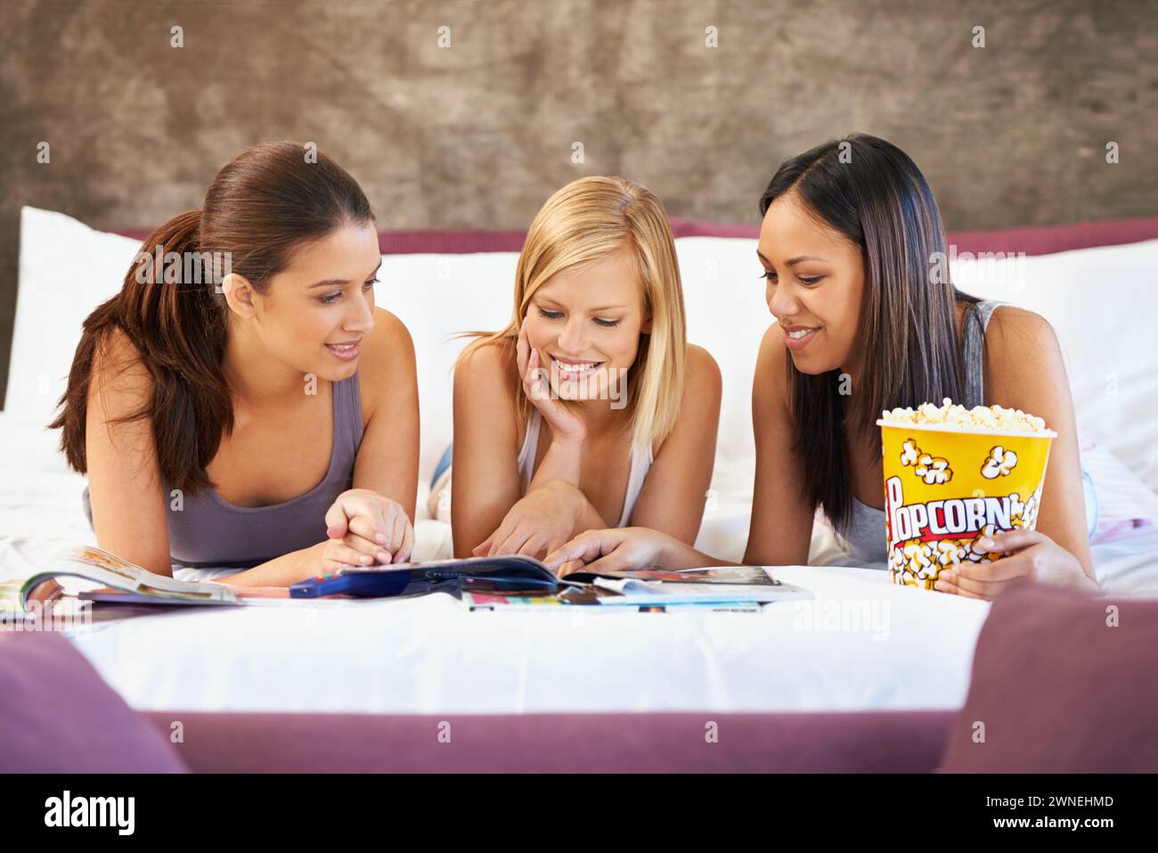 Smile, friends and women reading magazine in bedroom, food and eating popcorn snack together at party. Happy, girls and group with journal in home for Stock Photo