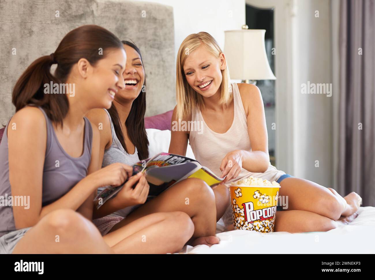 Friends, popcorn and reading magazine on bed with laughing, discussion and girls night in home with pyjamas. Women, snack and happiness with paper in Stock Photo