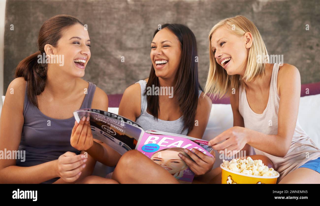 Friends, laughing and magazine on bed with happiness, girls night and entertainment in home with popcorn. Women, paper and excited with snack in house Stock Photo
