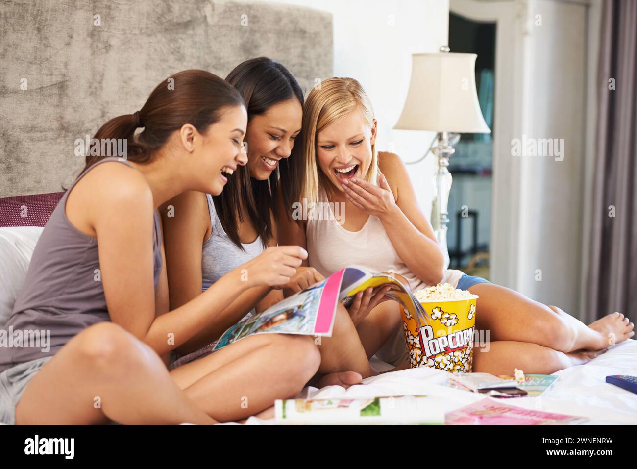 Friends, relax and magazine on bed with laughing, happiness and media entertainment in home. Women, excited and paper with popcorn for reading Stock Photo