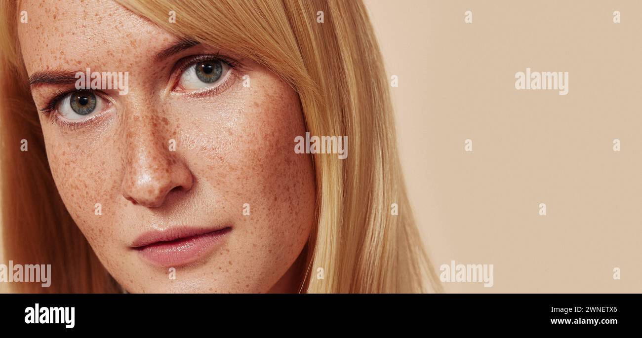 Close-up studio shot of a young beautiful female with naturally smooth skin. Cropped studio shot of woman with freckles looking at camera over beige b Stock Photo