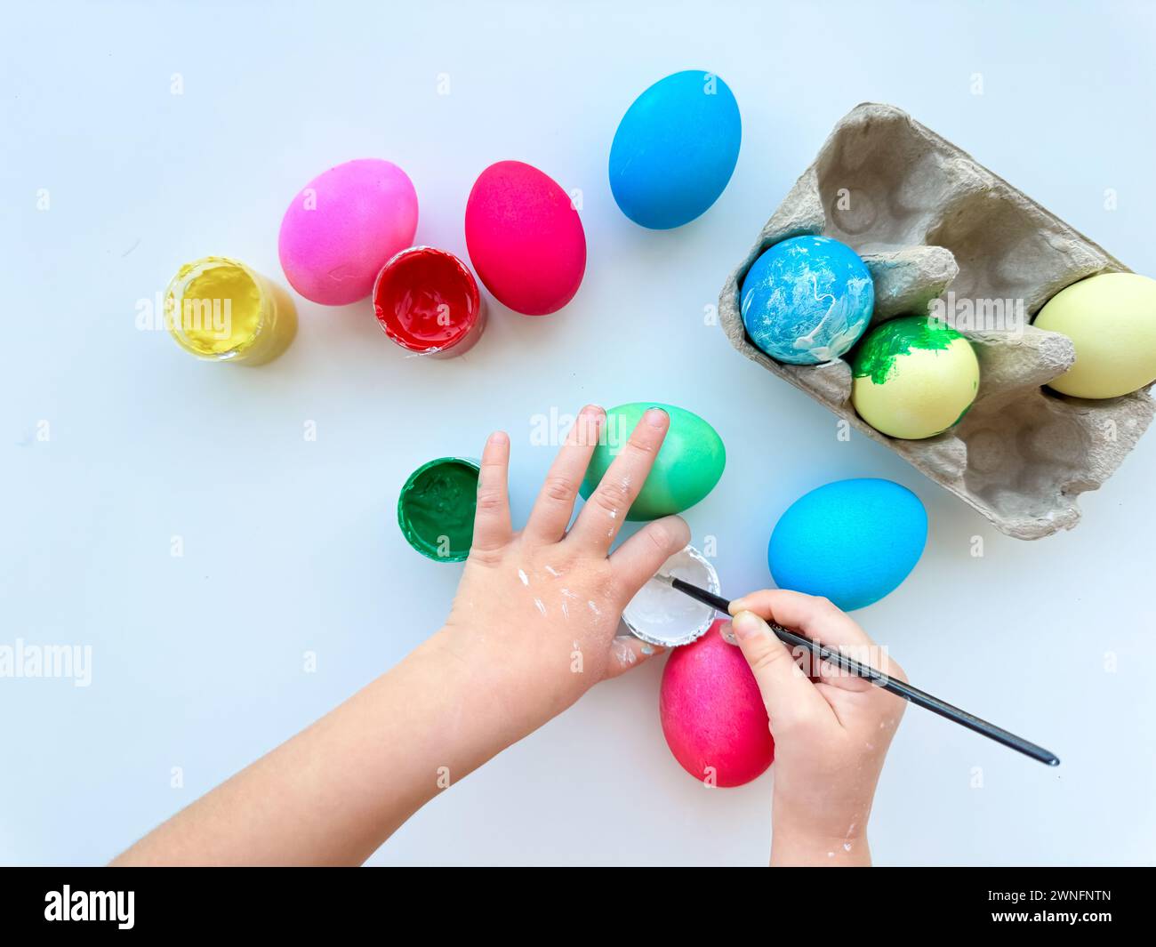 Childrens hands paint Easter eggs with brush surrounded by colorful eggs and jars of paints on white table, creative holiday activity. Stock Photo