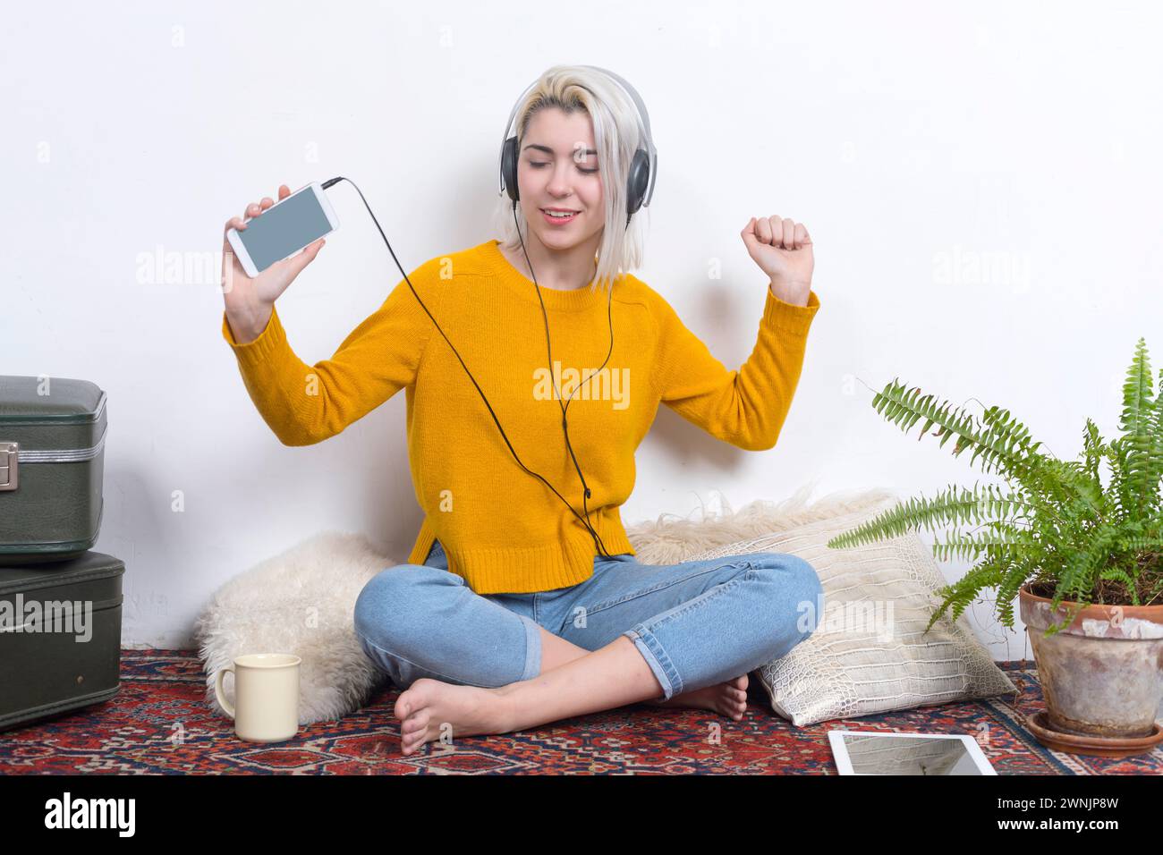 A happy woman with headphones dances while showing a smartphone, radiating enjoyment and energy in a homely, vintage-decorated space with plants. Stock Photo