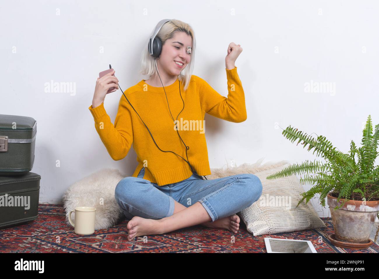 A happy woman with headphones dances while showing a smartphone, radiating enjoyment and energy in a homely, vintage-decorated space with plants. Stock Photo