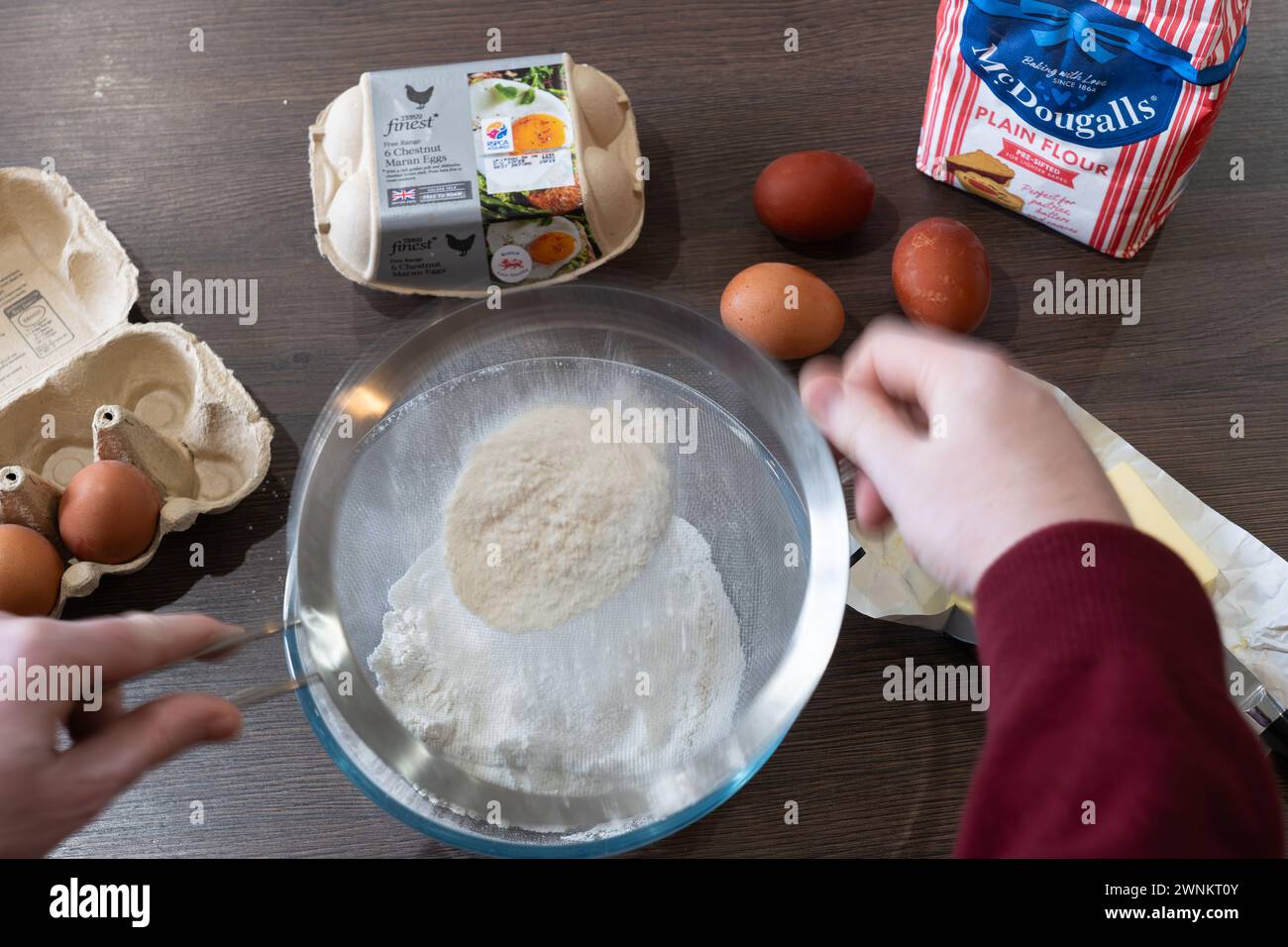 A man's hands holding a sieve and sifting flour, with ingredients for baking a cake on a kitchen worktop - butter, eggs and flour. UK Stock Photo