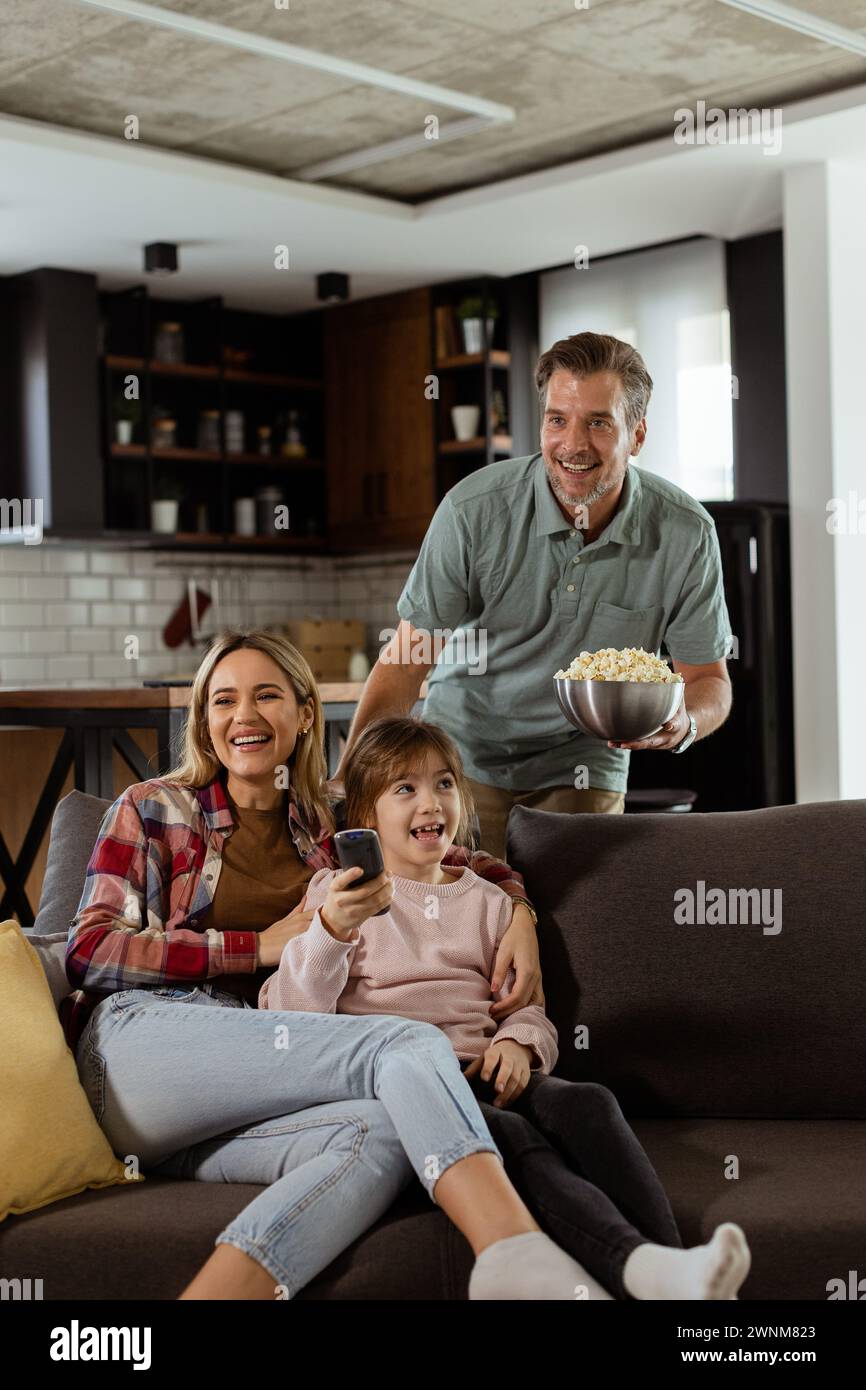 A family of three is comfortably nestled on a couch, their faces reflecting excitement and attentiveness as they share a bowl of popcorn during a susp Stock Photo