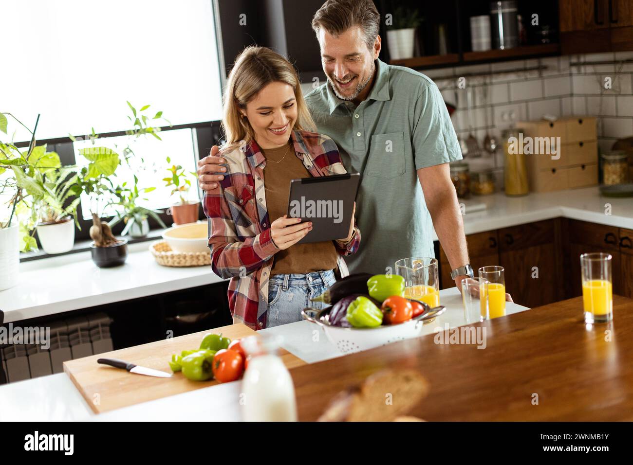 A cheerful couple stands in a well-lit kitchen, engrossed in a digital tablet among fresh ingredients Stock Photo