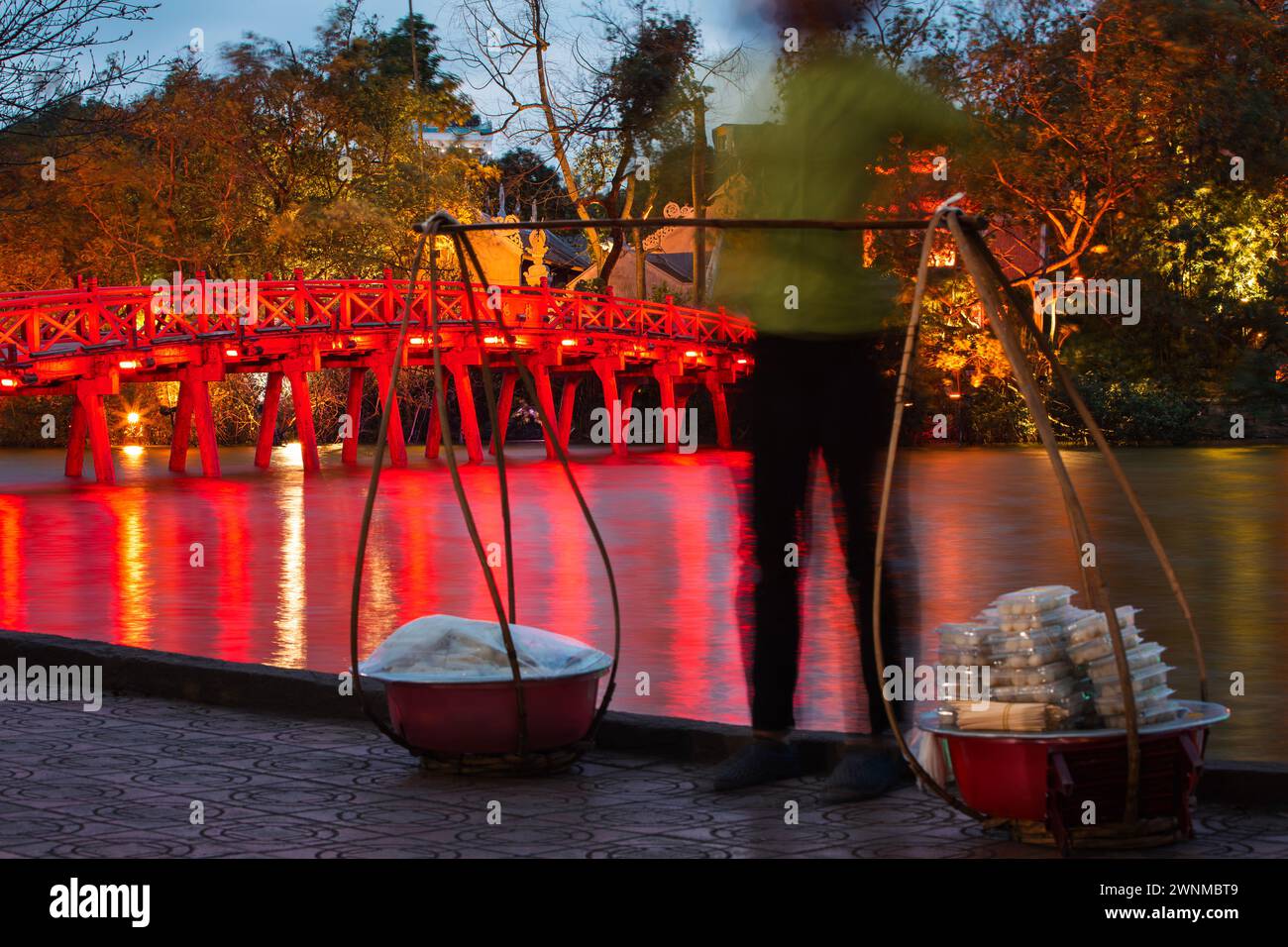 Vietnamese street food vendor in blurred motion against illuminated red Huc Bridge on Hoan Kiem lake in Hanoi, Vietnam. Stock Photo