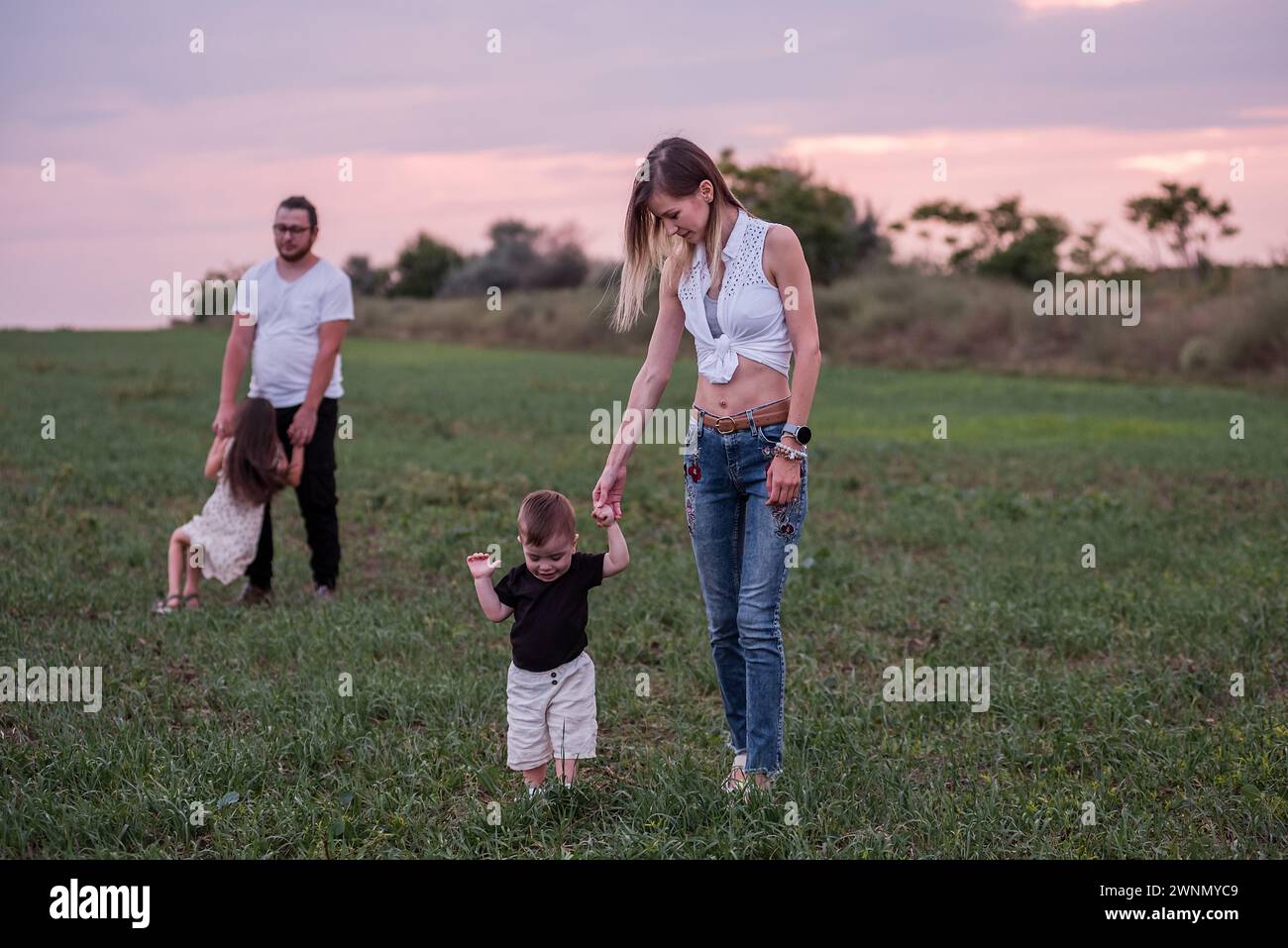 Woman helps toddler walk in a grassy field at sunset, with man and child in the background, capturing a casual, loving diversity family moment in the Stock Photo