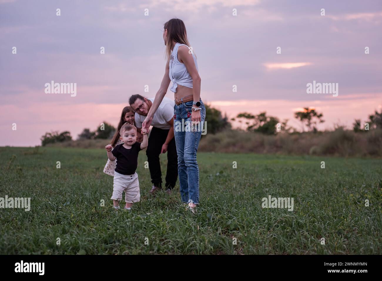 Woman helps toddler walk in a grassy field at sunset, with man and child in the background, capturing a casual, loving diversity family moment in the Stock Photo