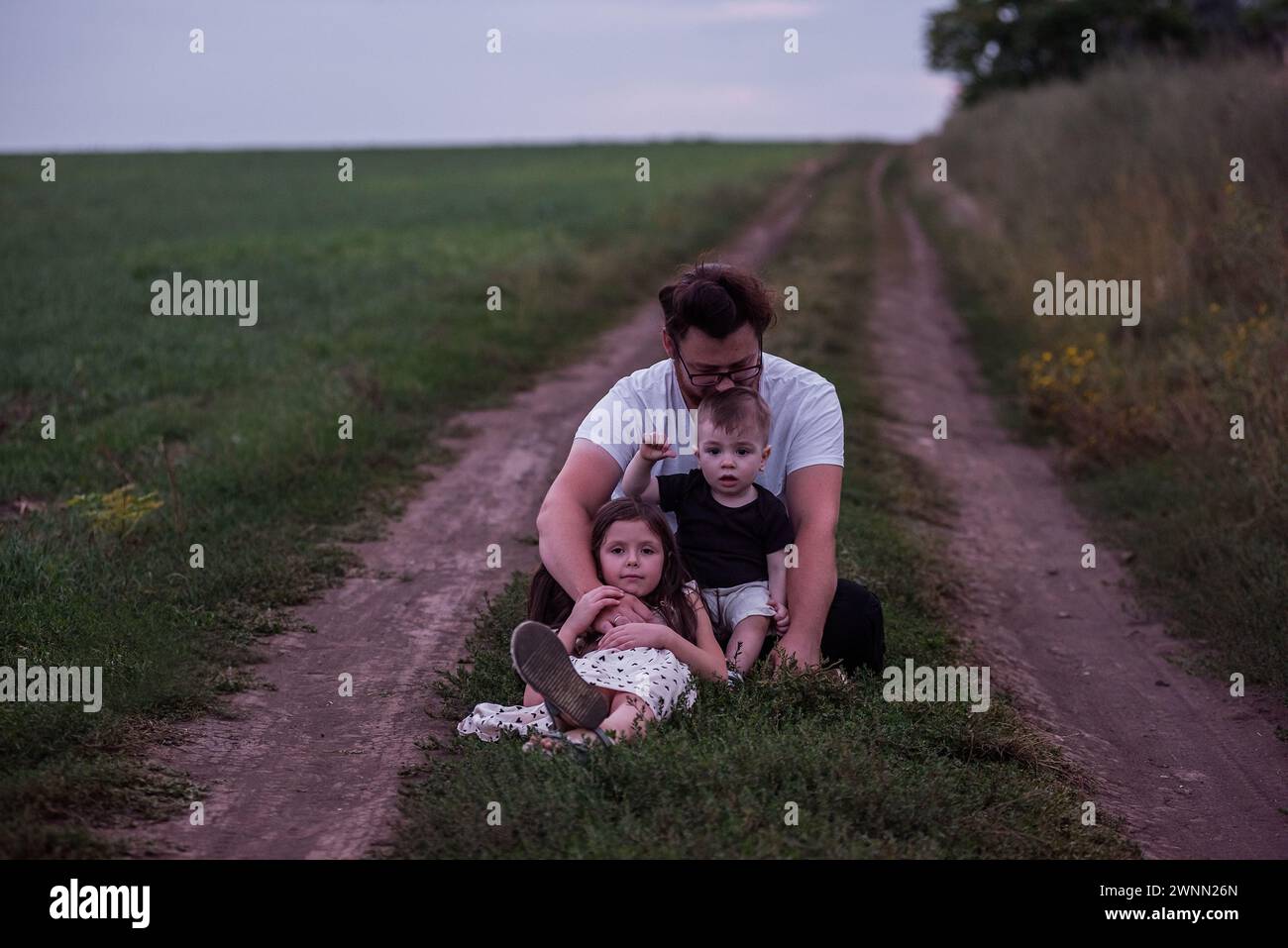 In the dimming light of dusk, father sits on rural trail, his children nestled in his arms, picture of protective love, quiet contemplation. Little gi Stock Photo