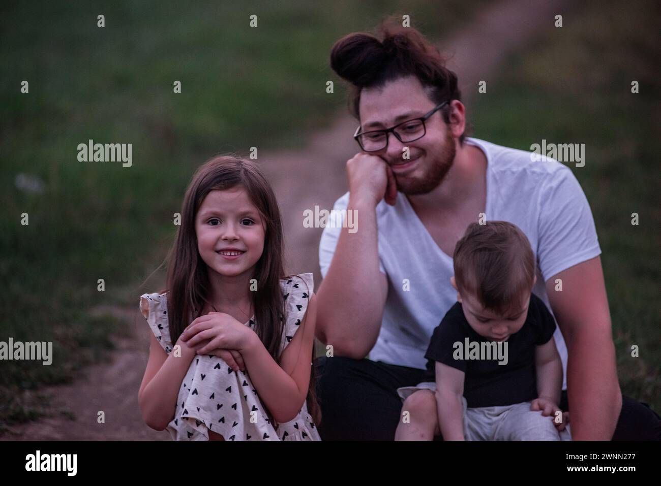 In the dimming light of dusk, father sits on rural trail, his children nestled in his arms, picture of protective love, quiet contemplation. Little gi Stock Photo