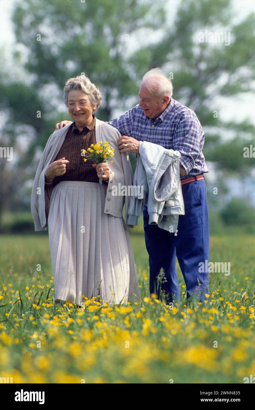 romantic senior couple in love stand on flowers man care about his wife balade outdoor trees in background Stock Photo