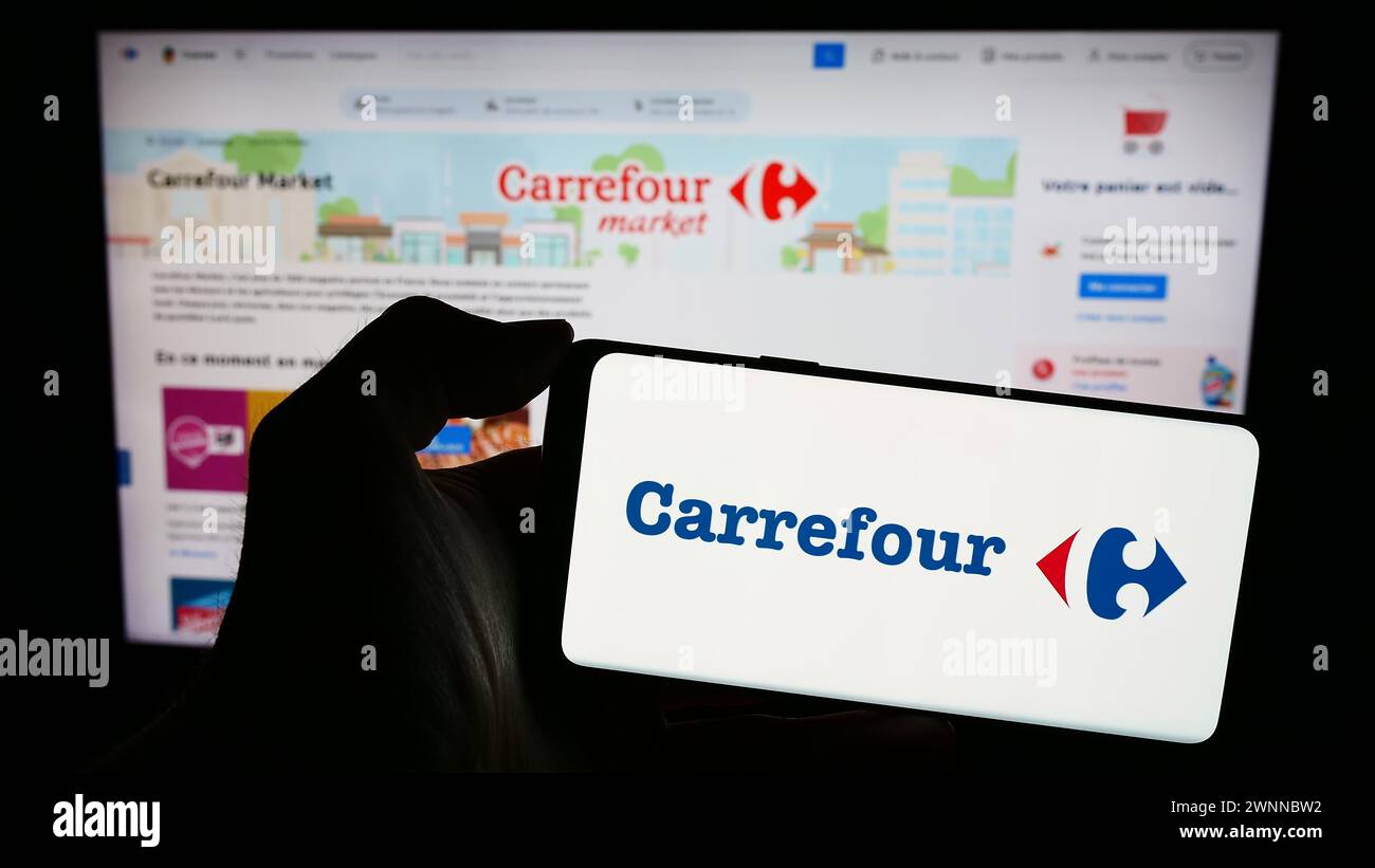 Person holding smartphone with logo of French retail company Carrefour S.A. in front of website. Focus on phone display. Stock Photo