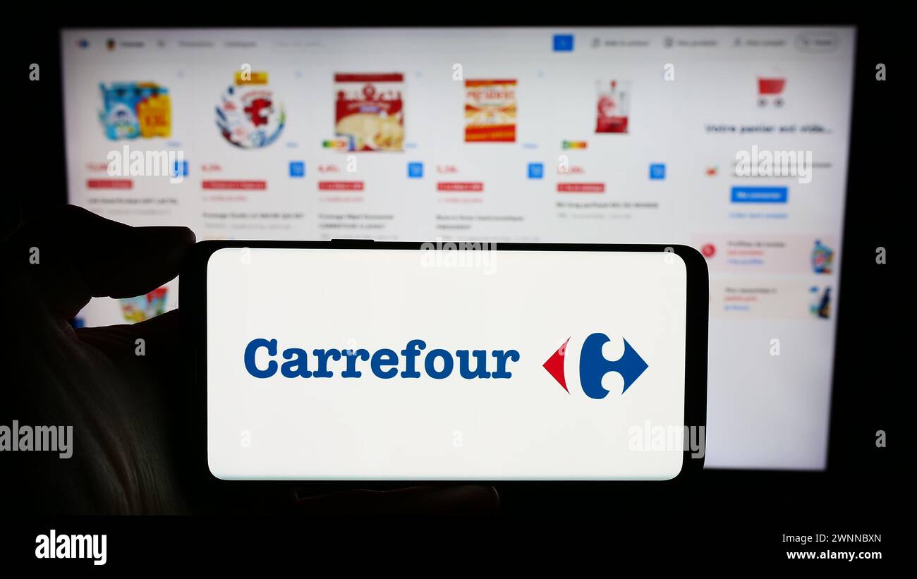 Person holding cellphone with logo of French retail company Carrefour SA in front of business webpage. Focus on phone display. Stock Photo
