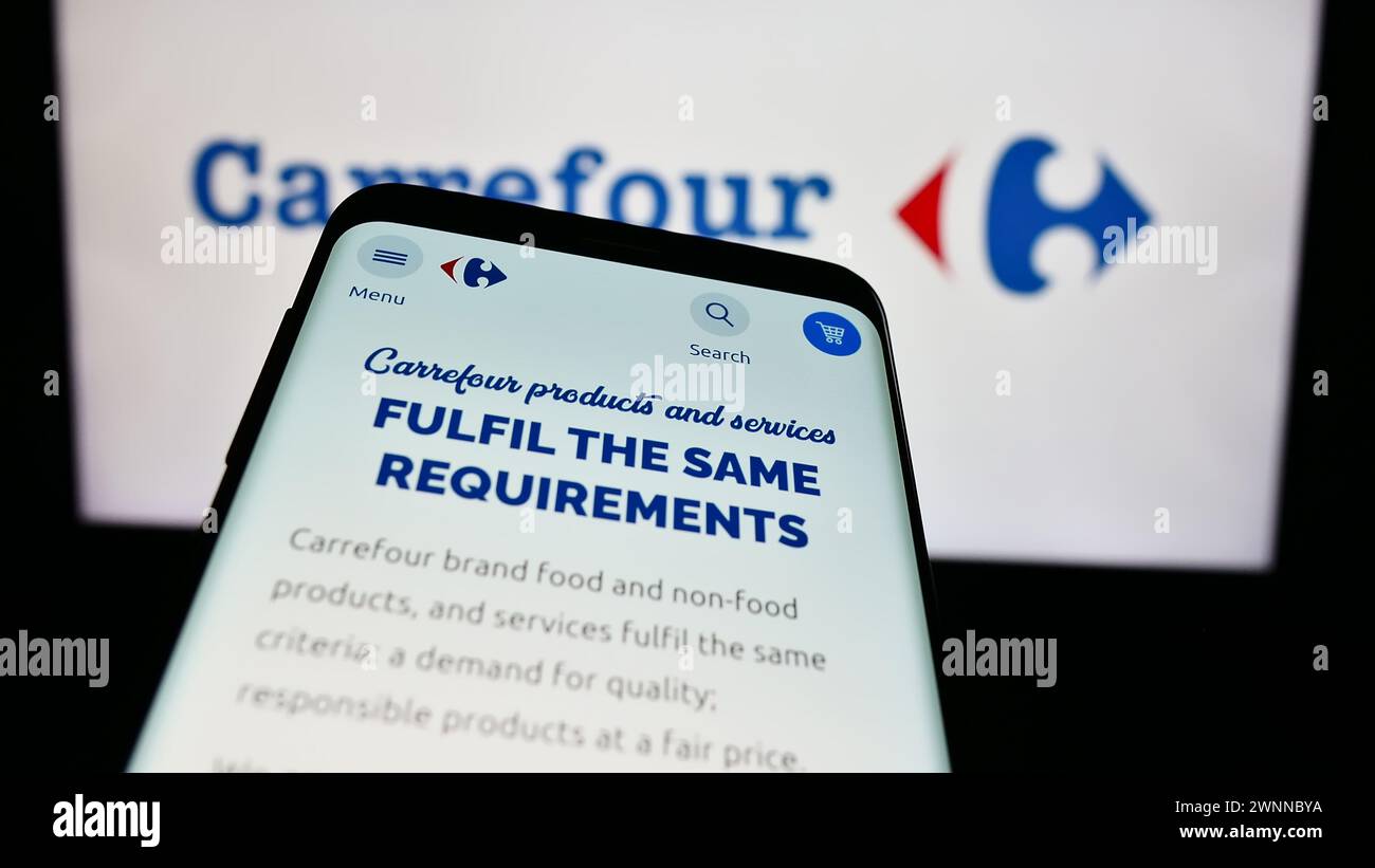Mobile phone with website of French retail company Carrefour S.A. in front of business logo. Focus on top-left of phone display. Stock Photo