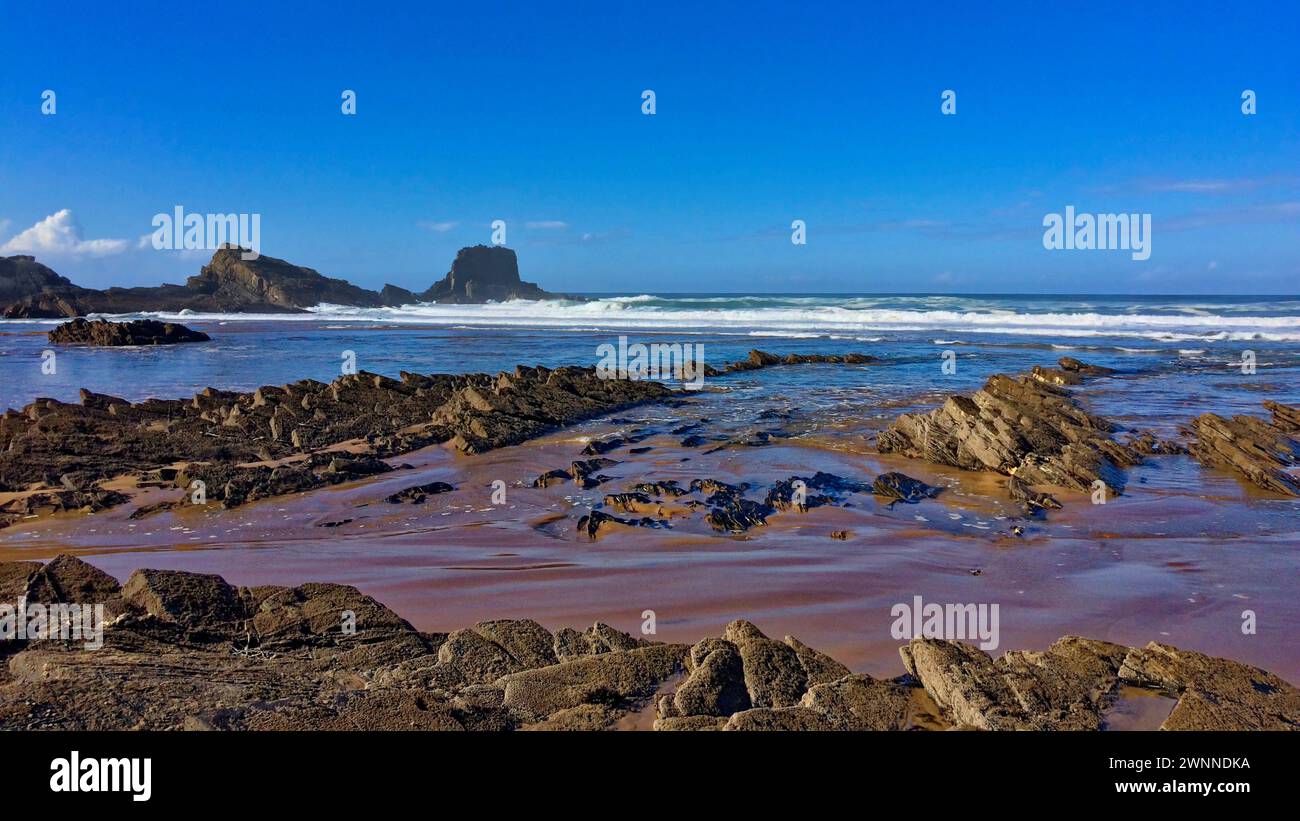 A sunny day at the beach showcasing layered rocks, waves, and an imposing cliff under a partly cloudy sky. Stock Photo