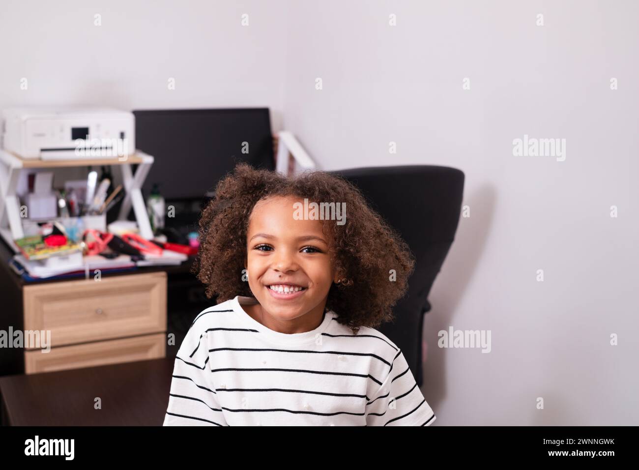 A cute and adorable black young girl with curly hair is smiling on a work or study desk on the background. Themes of home schooled kids, learning and Stock Photo