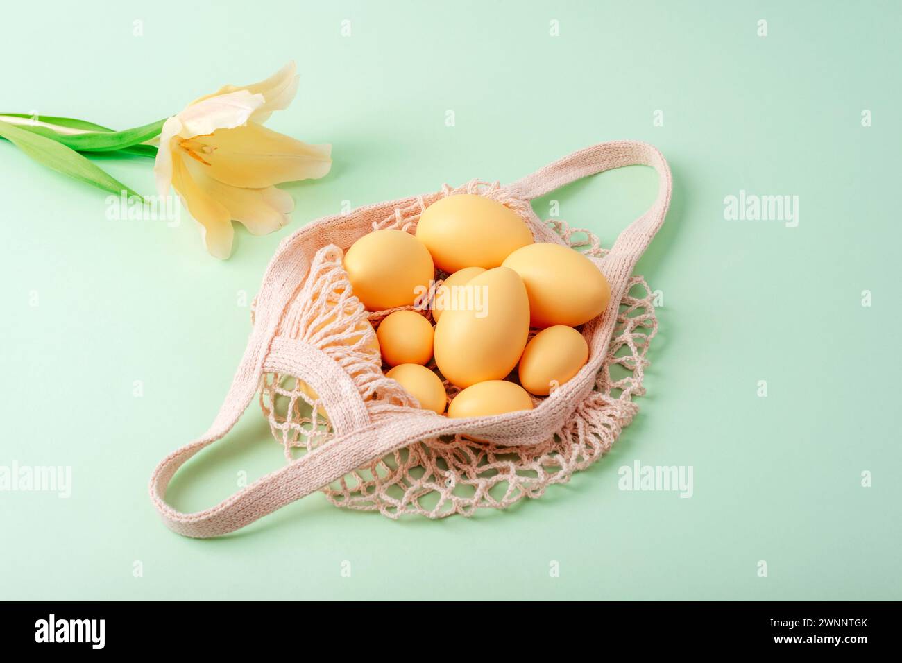 String bag with Easter eggs and yellow tulip on light green background. Top view, flat lay. Stock Photo