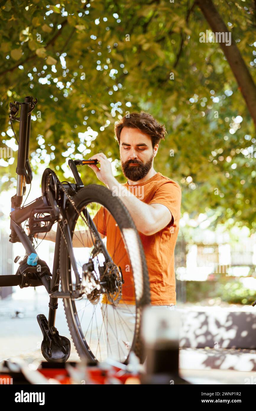 Healthy sports-loving man securing bike wheel for outdoor leisure cycling. Active young caucasian male cyclist doing yearly maintenance and adjusting bicycle tire in yard during the summer. Stock Photo