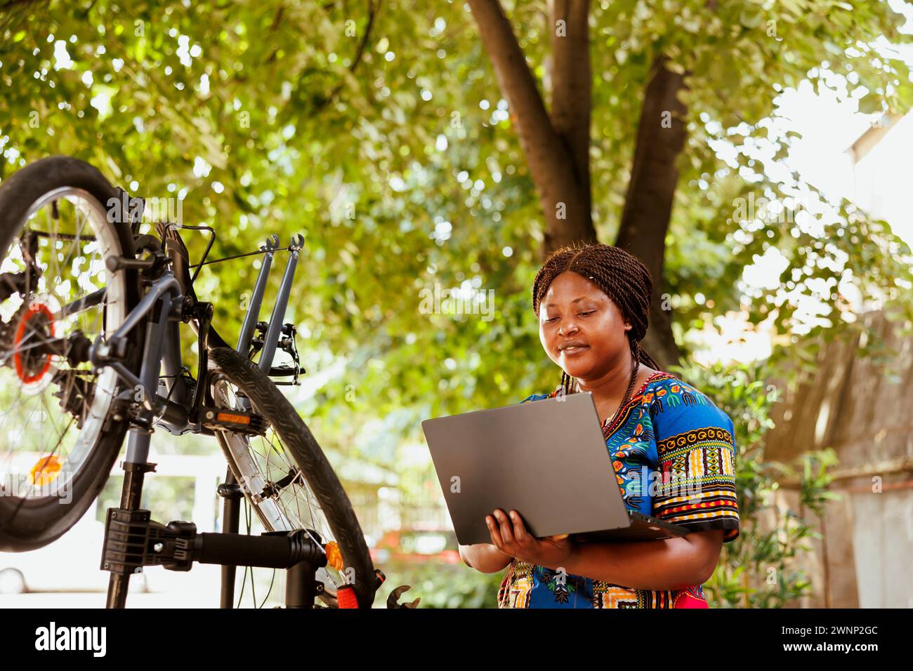 Healthy dedicated african american woman searching on laptop to fix broken modern bicycle. Active female cyclist ensures bike components are secure for summer outdoor leisure cycling. Stock Photo