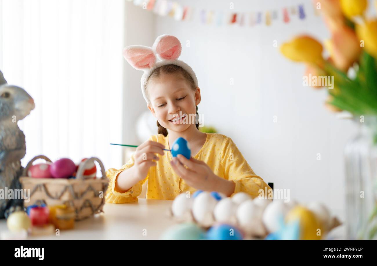 Beautiful child painting eggs. Happy family preparing for Easter. Cute little girl is wearing bunny ears. Stock Photo