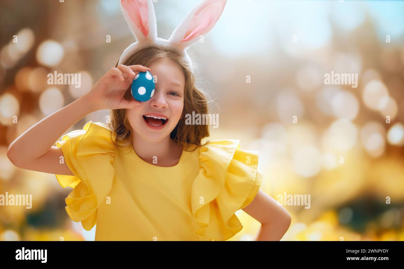 Beautiful child with painting eggs outdoors. Happy family celebrating Easter in nature. Cute little girl is wearing bunny ears. Stock Photo
