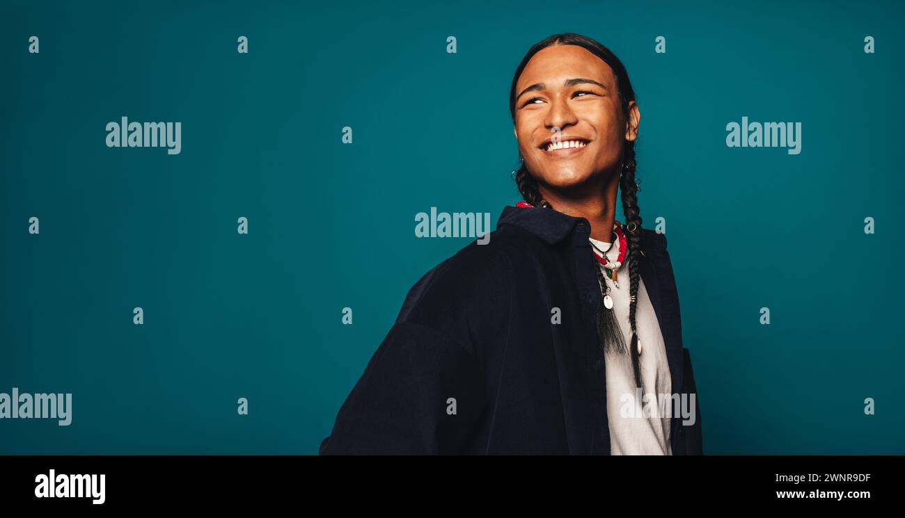 Confident, stylish young man with braided hair stands on a blue background. He wears casual clothing and a necklace, smiling happily while looking awa Stock Photo