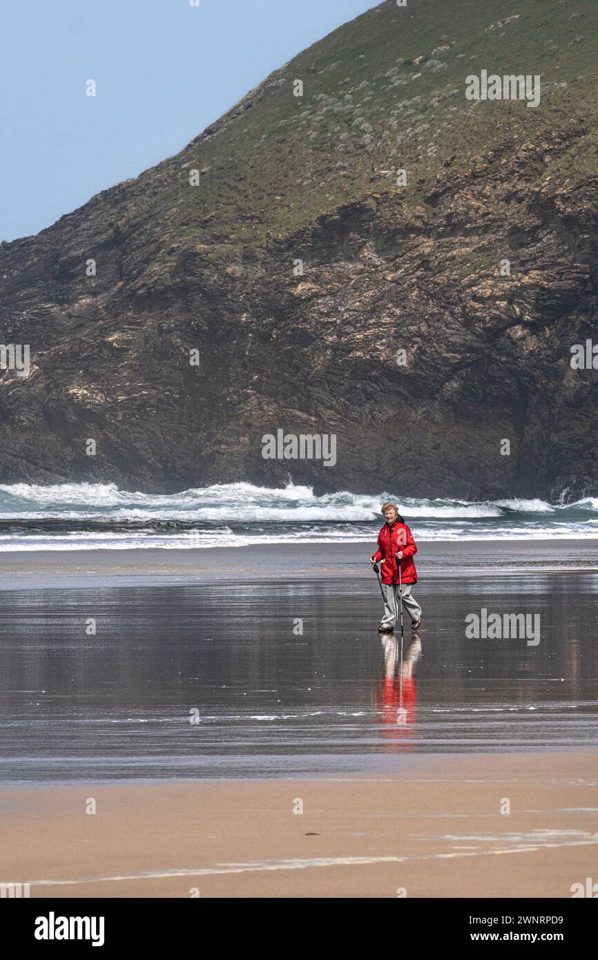 A mature female wearing a bright red coat and using walking poles hiking sticks walking on the shore at Mawgan Porth beach in Cornwall in the UK. Stock Photo